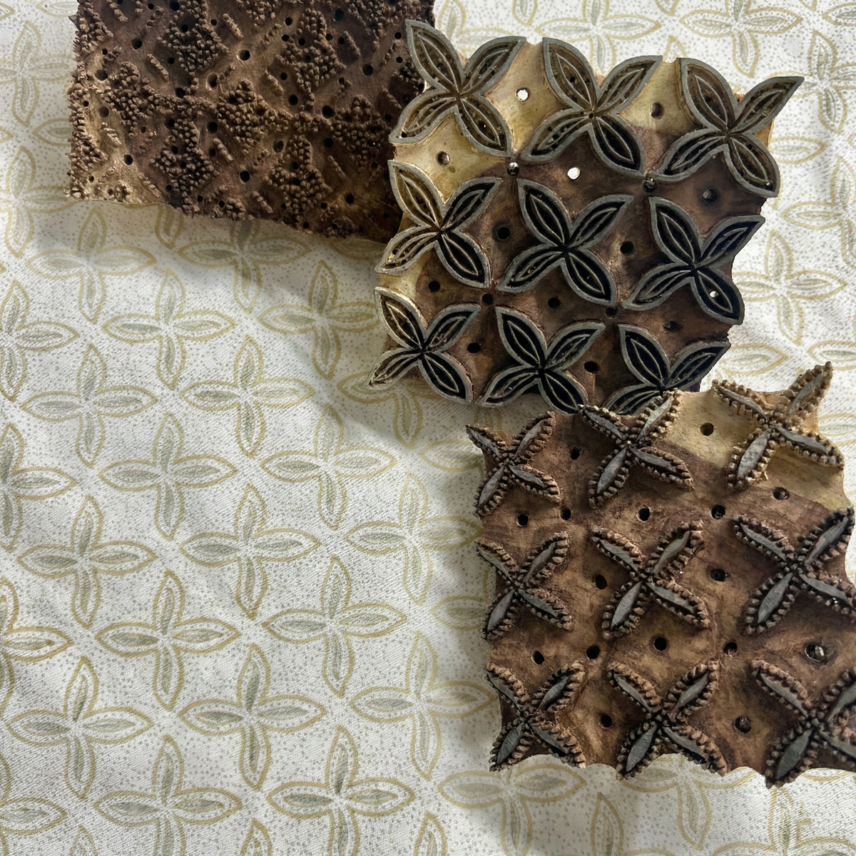 A close-up view of three intricately carved wooden blocks resting on patterned fabric, showcasing the traditional hand-block printing process used for BaliplumeriaBreezeThrowPillowCover-homewayliving.jpg. The blocks feature a delicate floral-inspired geometric design that, when stamped, creates the airy soft blue and earthy tan plumeria motifs on the crisp white textile. This artisanal process highlights the beautiful craftsmanship that perfectly adds a light, cozy modern rustic Canadian farmhouse aesthetic