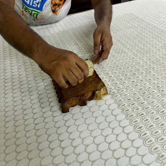 A close-up view of a skilled artisan's hands pressing a large carved wooden block onto natural fabric to create a delicate, repeating pale blue and beige scalloped arch pattern. This behind-the-scenes look highlights the traditional hand-block printing process used to craft our modern Canadian rustic home textiles.