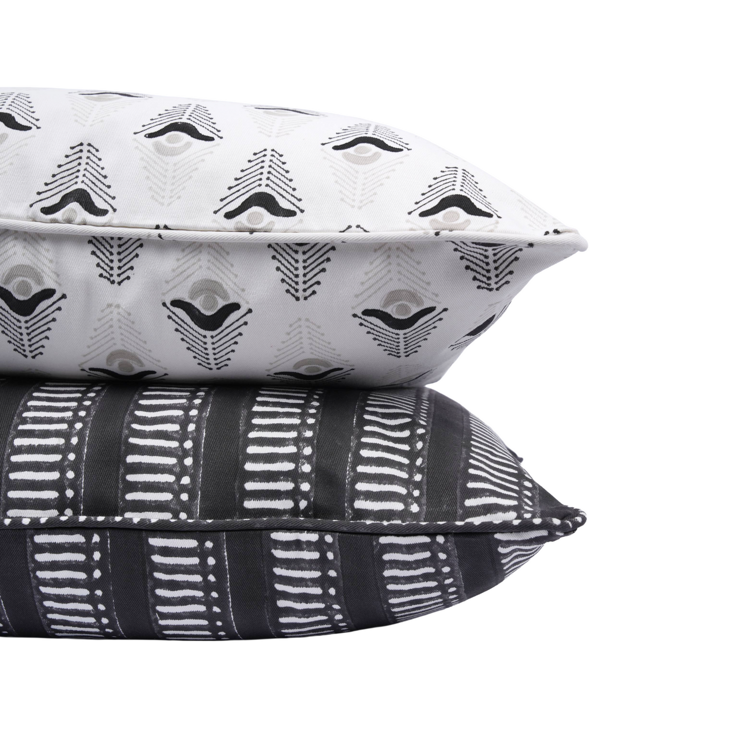 A close-up side view of two stacked hand-block printed cushions on a crisp white background. The top pillow features the white "Evia Charcoal" design with its modern abstract geometric motif, resting on a bold black cushion with a white horizontal dashed stripe pattern. Neatly finished piped edges highlight the quality of these handcrafted textiles, perfectly adding a bold yet cozy modern rustic Canadian farmhouse aesthetic to your home decor.