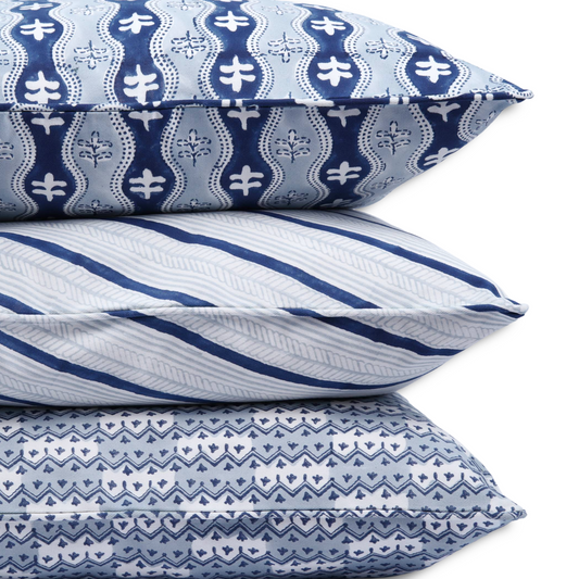A close-up of three stacked hand-block printed cushions on a white background. The top pillow features a blue wavy floral design, resting on the Serere Frost indigo striped cushion, supported by the Munnar Frost blue zig-zag pattern. These piped handcrafted textiles add a cozy modern rustic Canadian farmhouse aesthetic to your home decor.