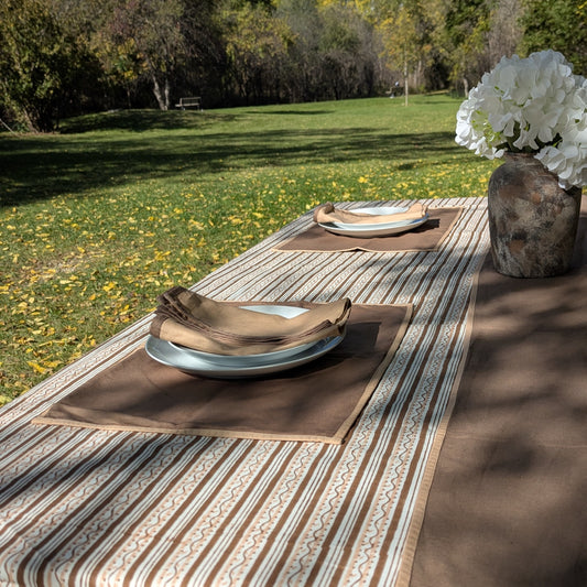 A styled outdoor dining table setup in a grassy park, featuring a striped beige and brown table runner, matching placemats, folded tan napkins, white plates, and a rustic vase with white flowers.