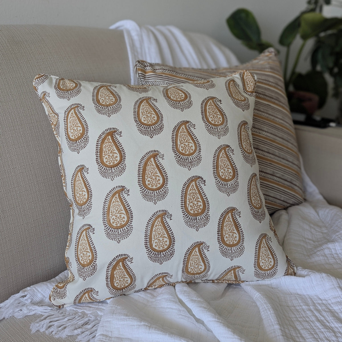 A close-up of a hand-block printed square cushion featuring an earthy brown and warm ochre paisley pattern on a cream background. Resting on a neutral textured sofa, this handcrafted textile perfectly adds a cozy modern rustic Canadian farmhouse aesthetic to your living room decor.