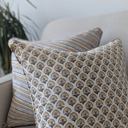 A close-up of two hand-block printed cushions resting on a neutral sofa. The foreground features a warm ochre and earthy brown geometric arch pattern, perfectly layered in front of a coordinating diagonal striped cushion. These handcrafted textiles effortlessly add a cozy modern rustic Canadian farmhouse aesthetic to your living room decor.