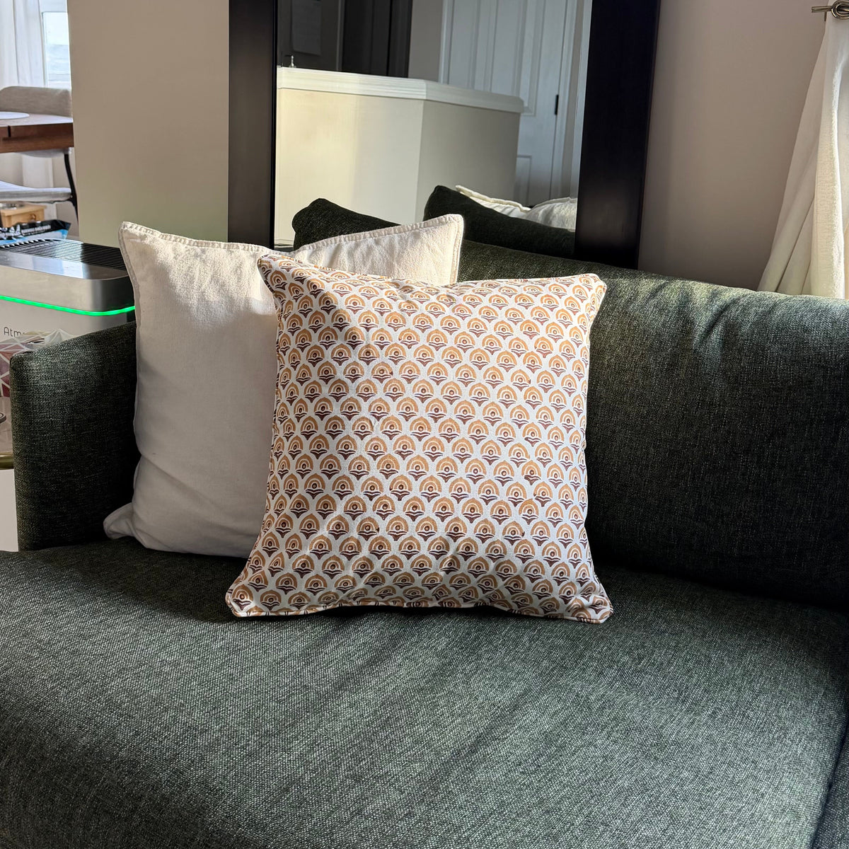 A green upholstered sofa in a living room setting, styled with two throw pillows: one solid white and one featuring a beige, orange, and blue geometric pattern. A large black-framed mirror is mounted on the wall behind the sofa.