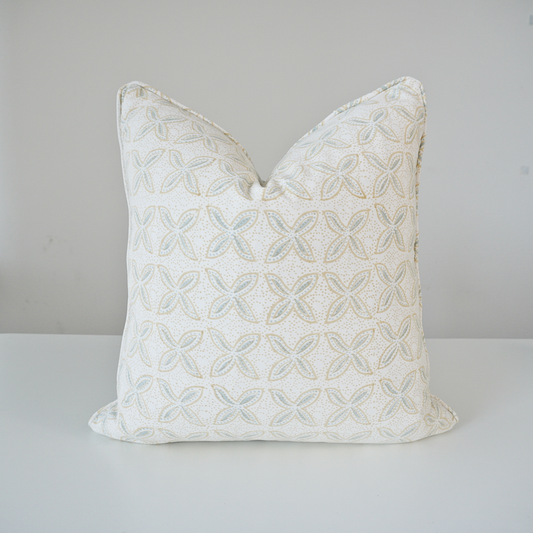 A square throw pillow with a textured, raised geometric floral pattern in light blue and beige on an off-white fabric background, displayed on a white surface against a neutral light gray wall.