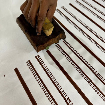Close-up view of an artisan using a hand-carved wooden block to print on white cotton fabric. Demonstrates the traditional hand block printing craft process of stamping a detailed stripe and dashed line pattern to create fine handmade throw pillow and cushion covers.