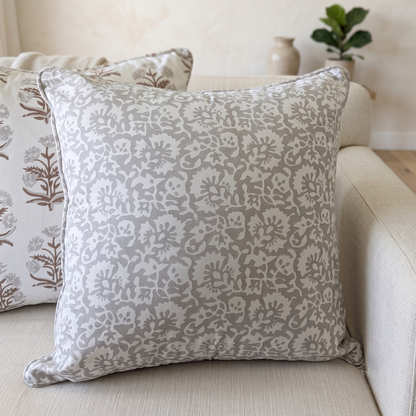 A close-up lifestyle shot of the Fiora Muse 100% cotton block printed pillow cover positioned on a light beige sofa. This artisan throw cushion features an intricate, dense floral botanical pattern in soft taupe and grey tones on a crisp white background, complemented by tailored piped edges. The clean, high-end design is showcased in a softly lit, modern home setting.