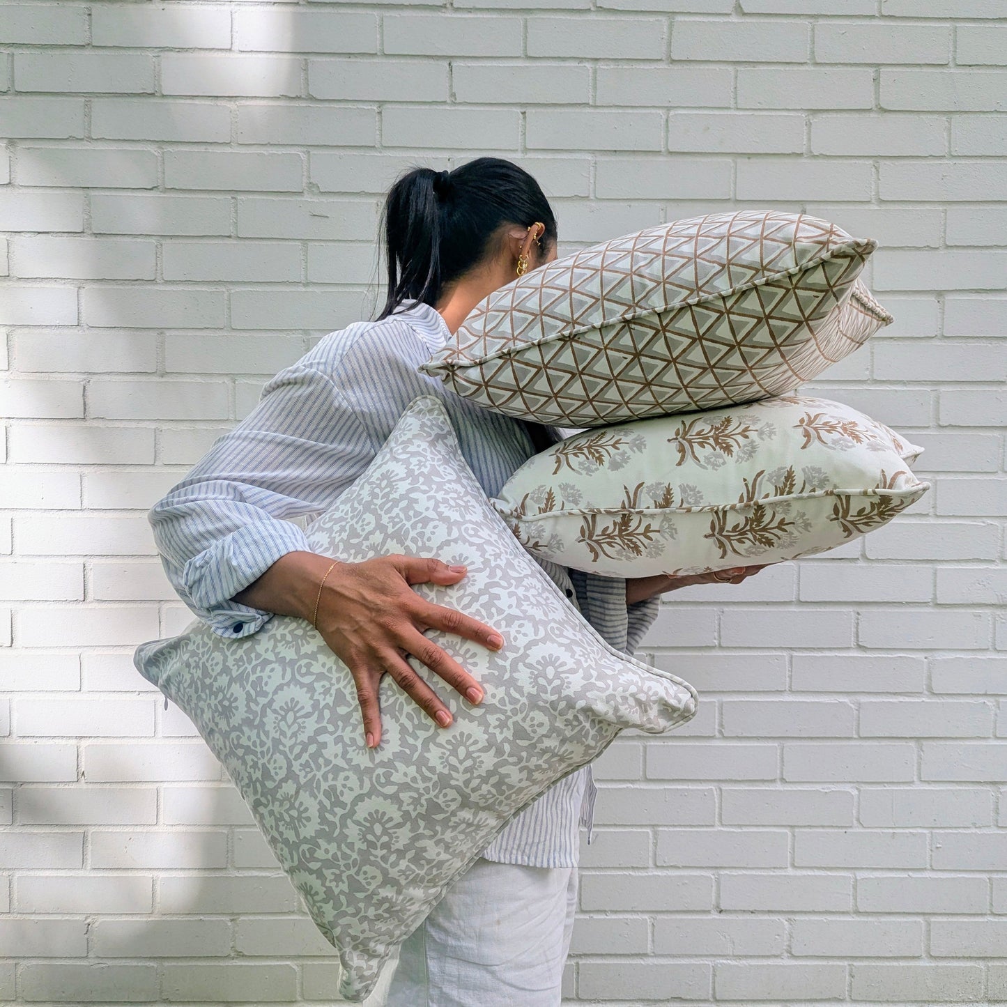 A lifestyle shot of someone holding three hand-block printed cushion covers against a sunny white brick wall. The bundle features brown geometric, floral sprig, and delicate beige motifs, adding a warm, modern rustic farmhouse aesthetic to any Canadian and USA home decor.