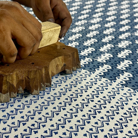 A close-up view of an artisan's hands firmly pressing a intricately carved wooden block onto fabric, demonstrating the traditional hand-block printing process. The textile reveals a striking blue and white geometric chevron pattern being stamped. This authentic craftsmanship highlights the quality of these handmade textiles, perfectly suited for adding a cozy modern rustic Canadian farmhouse aesthetic to your home decor.
