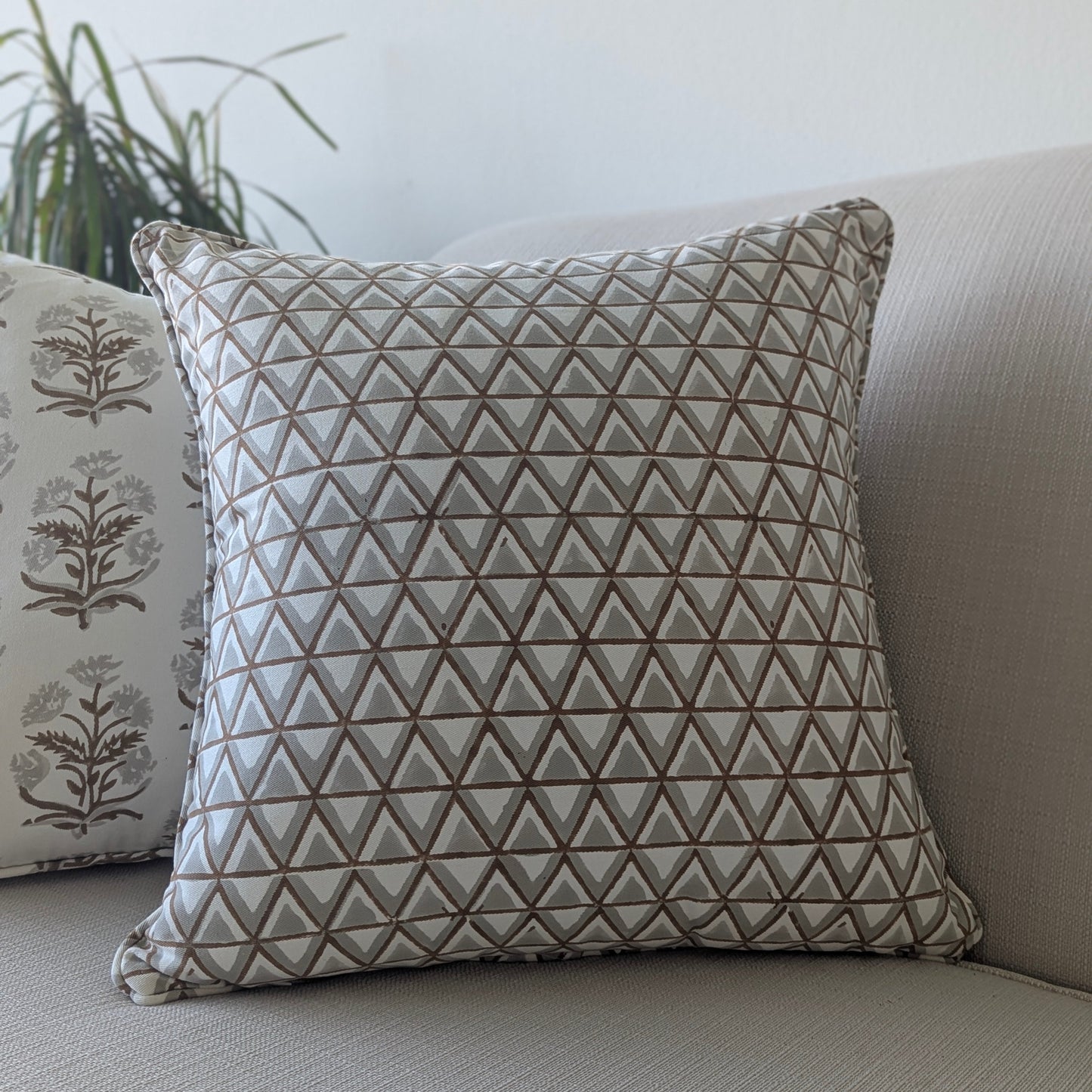 A close-up of a hand-block printed square cushion featuring an earthy tan and soft grey geometric triangle pattern on a cream background. Resting on a neutral textured sofa beside a coordinating botanical floral pillow, this handcrafted textile perfectly adds a cozy modern rustic Canadian farmhouse aesthetic to your living room decor.