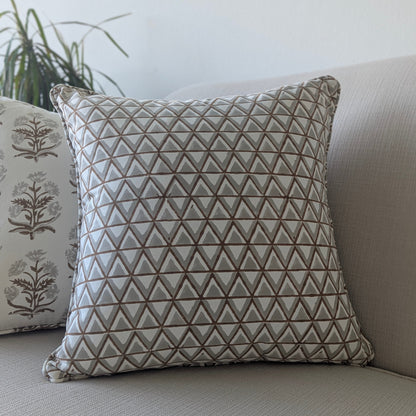 A close-up of a hand-block printed square cushion featuring an earthy tan and soft grey geometric triangle pattern on a cream background. Resting on a neutral textured sofa beside a coordinating botanical floral pillow, this handcrafted textile perfectly adds a cozy modern rustic Canadian farmhouse aesthetic to your living room decor.