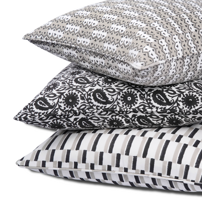 A vertical stack of three 100% cotton block printed pillow covers against a white background. The top cushion features the Munnar Stone grey and white chevron pattern. The middle displays the bold Siona Charcoal black and white floral paisley. The bottom is the Oslo Stone cover with a modern black and grey dashed geometric design. Each handmade artisan piece includes tailored piped edges.