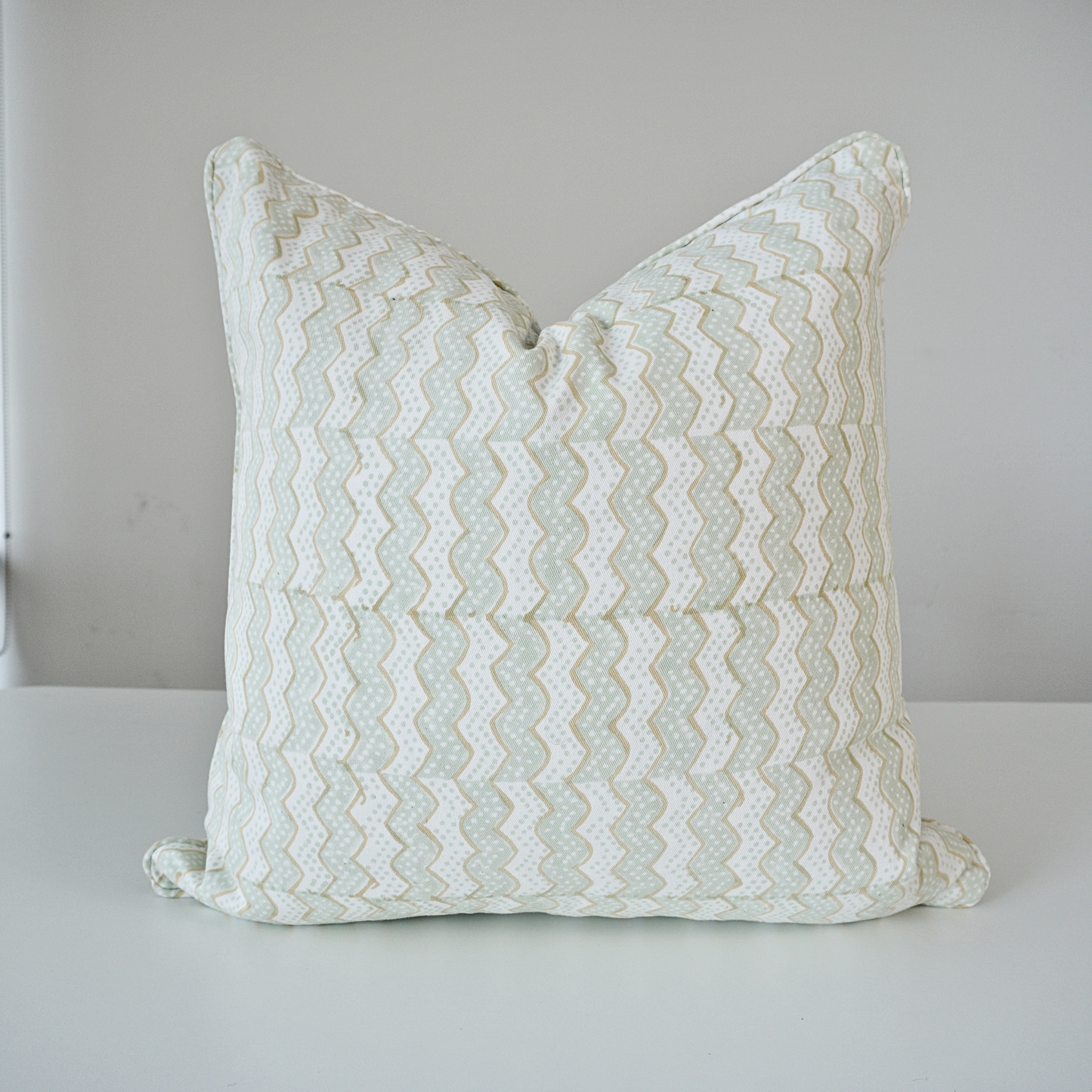 A square throw pillow with a light sage green and white chevron pattern, resting on a white surface against a neutral light gray background.