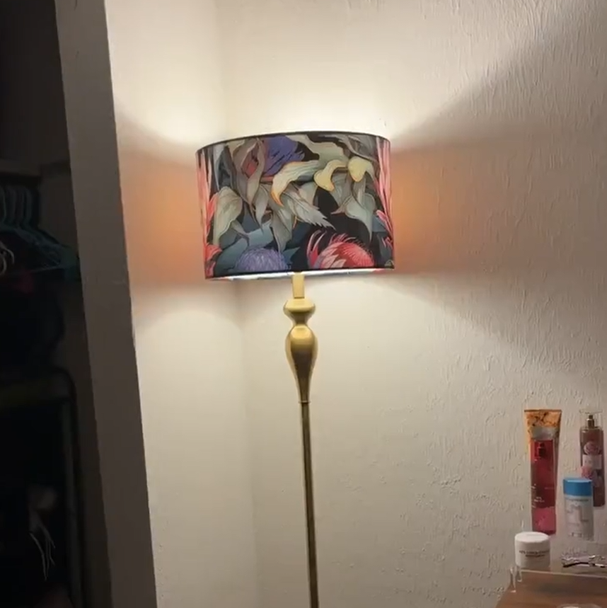 A floor lamp with a colorful floral-patterned drum shade and a gold-toned pole, standing in a room corner against a textured off-white wall.
