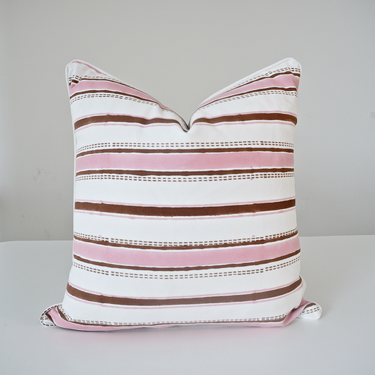 A square throw pillow with a horizontal striped pattern in white, pink, and brown, featuring dotted accent lines, resting on a white surface against a light gray background.