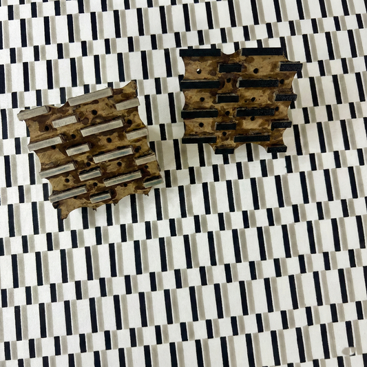 A close-up view of two carved wooden blocks resting on printed fabric, showcasing the traditional hand-block printing process for OsloStoneThrowPillowCover-homewayliving.jpg. The geometric rectangular bar design creates a striking staggered black, stone grey, and white broken stripe pattern. This artisanal process highlights the craftsmanship that perfectly adds a cozy modern rustic Canadian farmhouse aesthetic to your home decor.