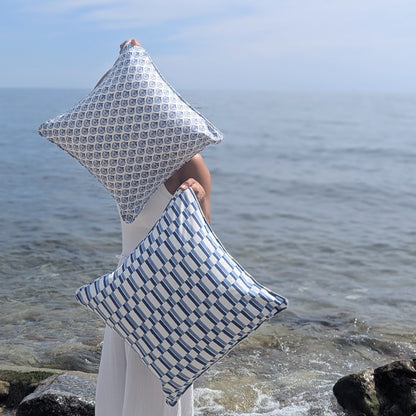 Two square throw pillows with blue and white geometric patterns are held by a person standing on a rocky shoreline with the ocean in the background.