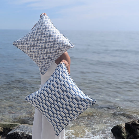 Two square throw pillows with blue and white geometric patterns are held by a person standing on a rocky shoreline with the ocean in the background.