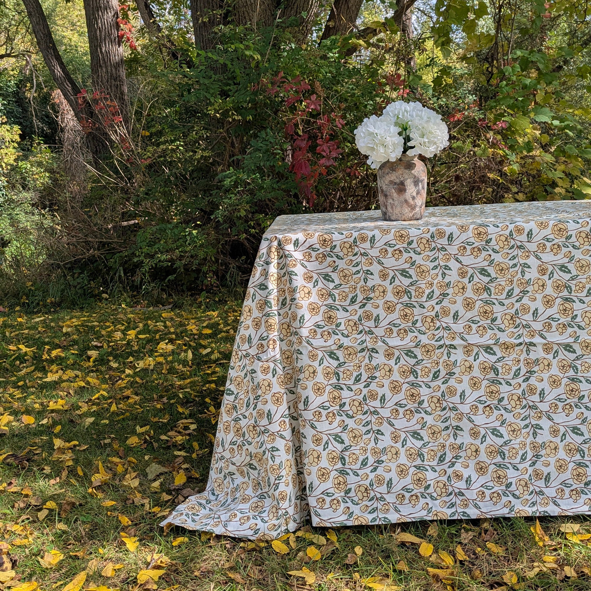 A rectangular table covered with a white tablecloth featuring a repeating yellow and green floral pattern, set outdoors on grass with fallen leaves and dense foliage in the background. A small vase with white flowers sits on the tabletop.