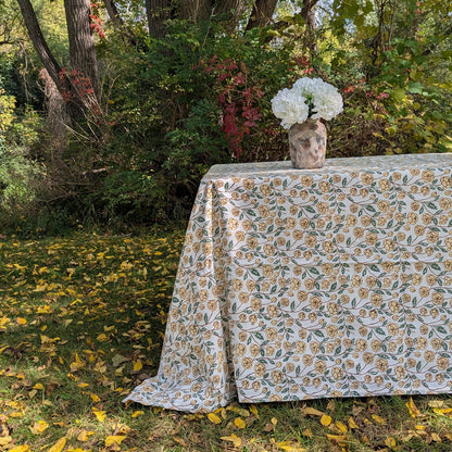 A rectangular table covered with a white tablecloth featuring a repeating yellow and green floral pattern, set outdoors on grass with fallen leaves and dense foliage in the background. A small vase with white flowers sits on the tabletop.