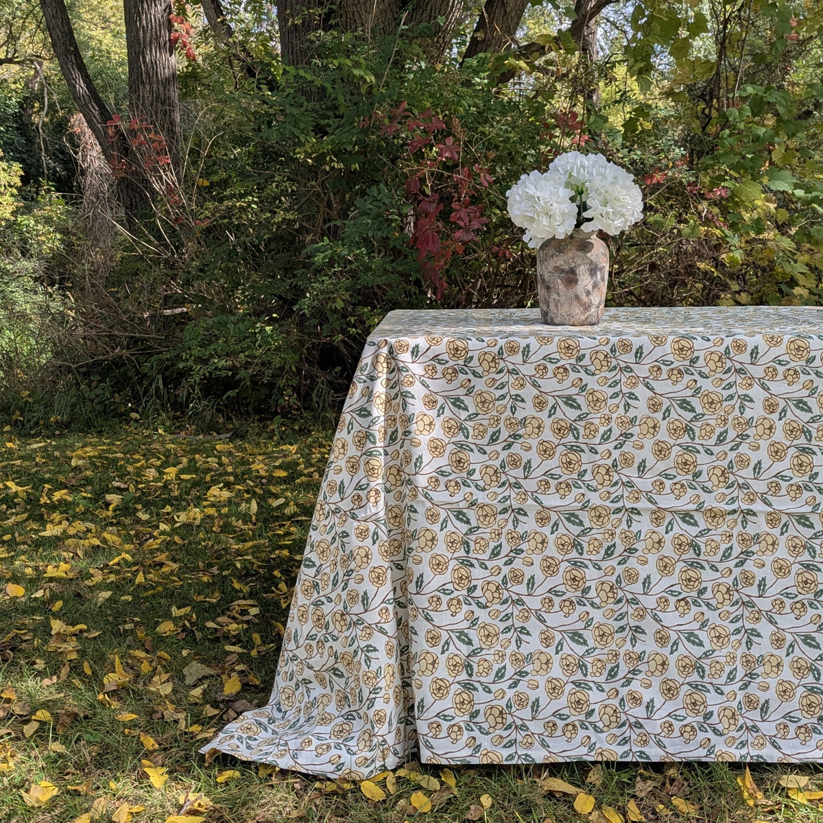 A rectangular tablecloth with a vintage floral pattern in muted yellow and green on an off-white background, draped over an outdoor table with a vase of white flowers, set against a backdrop of trees and fallen leaves.