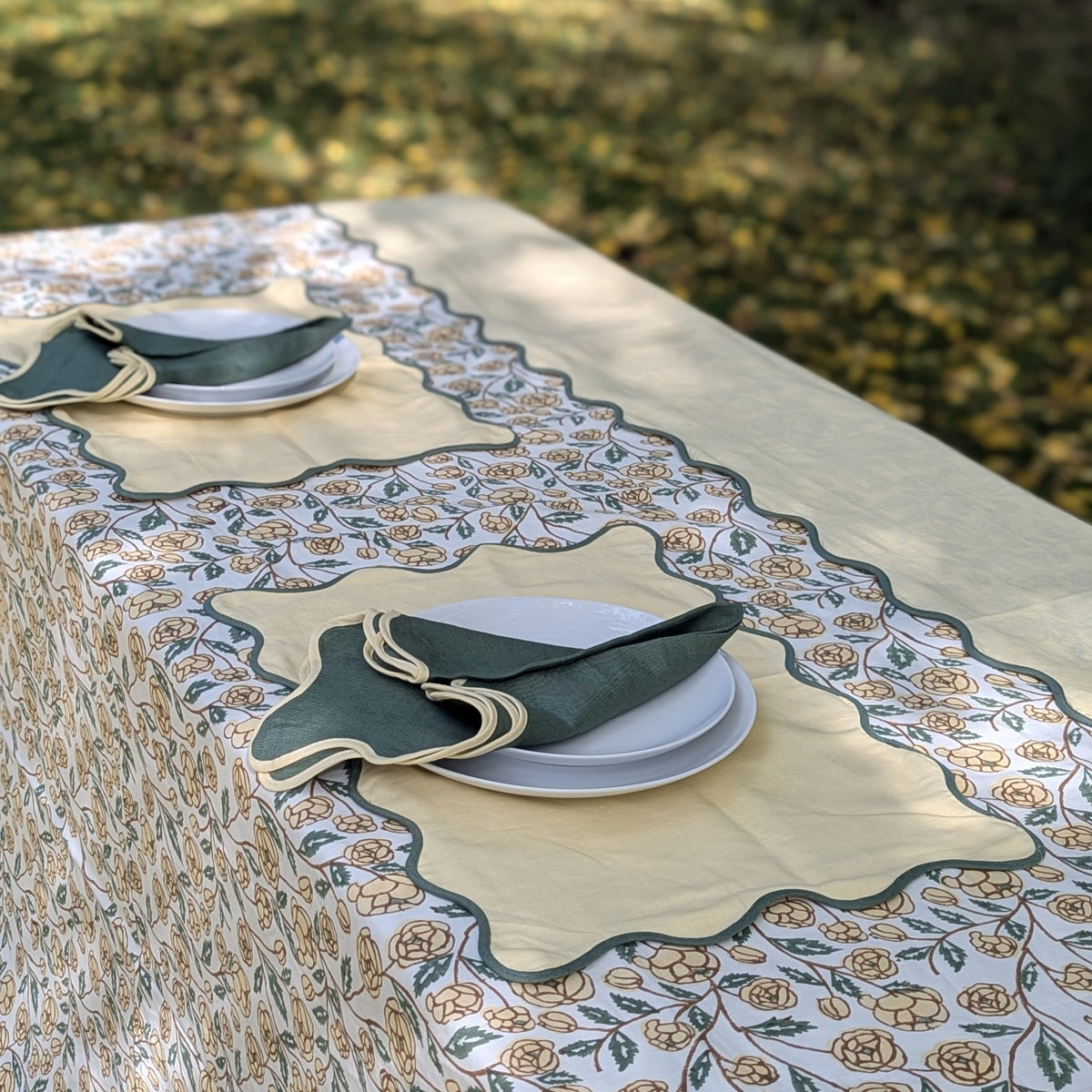 A rectangular outdoor table set with a cream tablecloth featuring a beige and green floral pattern, a matching scalloped runner, white plates, and folded dark green napkins. The background shows blurred green grass and foliage.