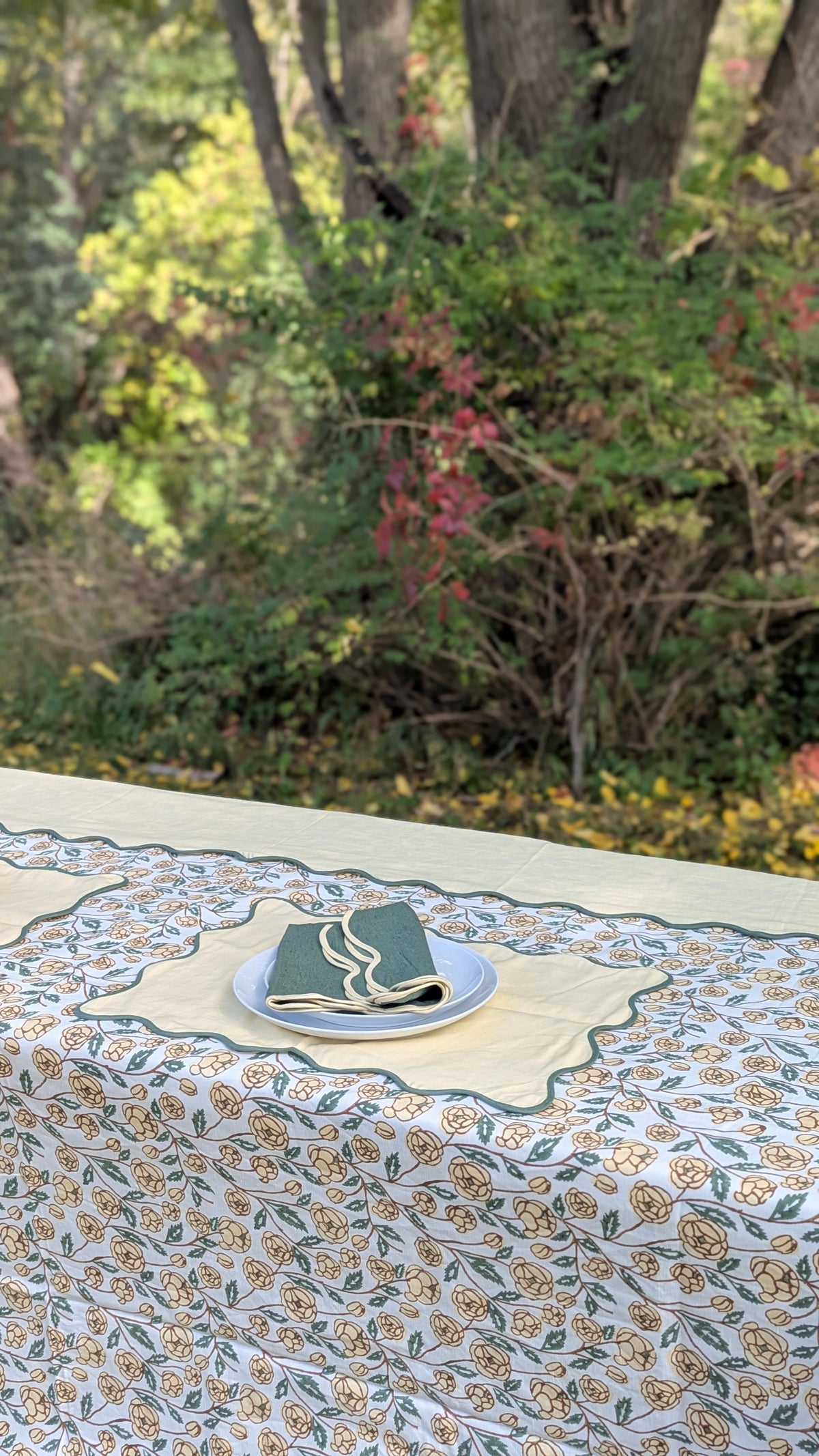 A close-up of a patterned tablecloth with a floral print in beige, green, and blue, styled with a folded green napkin on a blue plate on an outdoor table. The background is a soft-focus natural forest setting.