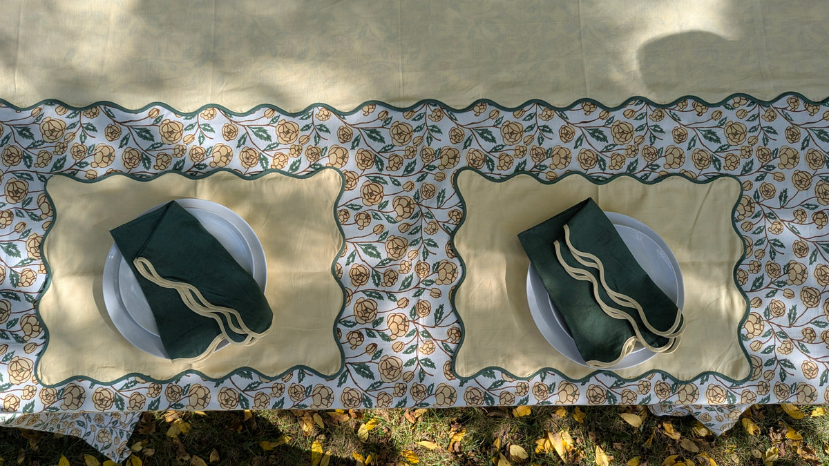 A top-down view of a table set with a beige tablecloth featuring a decorative floral border, two beige placemats, white plates, and dark green napkins with gold embroidered details.