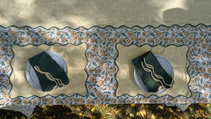 A top-down view of a table set with a beige tablecloth featuring a decorative floral border, two beige placemats, white plates, and dark green napkins with gold embroidered details.
