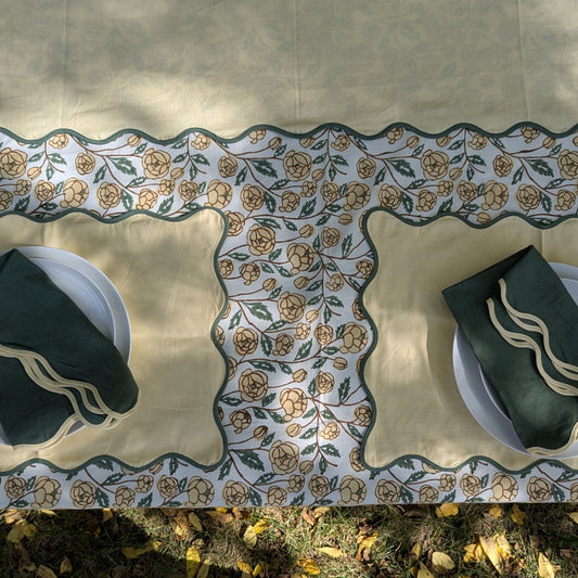 A rectangular tablecloth with a cream base and a central floral border pattern in tan and green, styled outdoors on a grassy ground with two place settings visible.