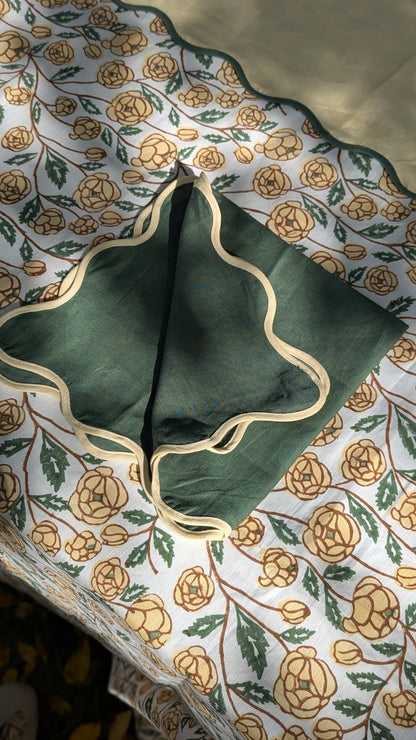 A dark green square fabric napkin with a scalloped cream border, laid flat on a white tablecloth patterned with yellow and green roses.