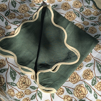 A dark green fabric pouch with a scalloped beige trim, laid flat on a white floral-patterned fabric background.