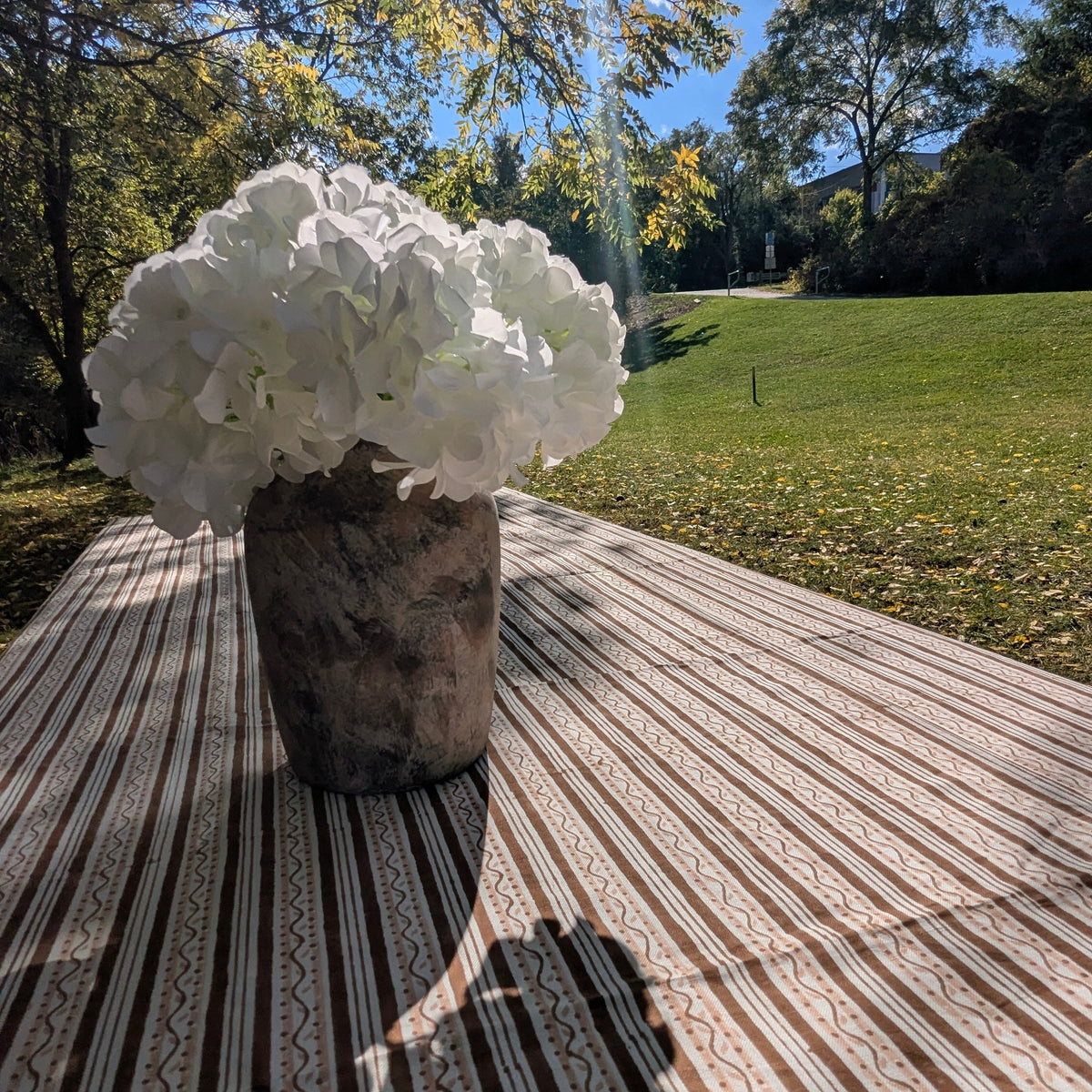 A rustic wooden vase filled with large white flowers sits on a striped beige and brown tablecloth outdoors on a wooden deck, with a sunny lawn, trees, and blue sky in the background.