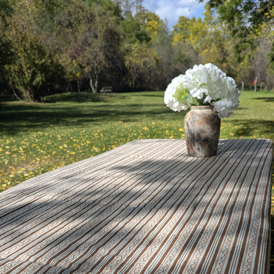 A rectangular outdoor dining table covered with a beige and brown striped tablecloth, set in a grassy park with a rustic vase of white flowers on top.