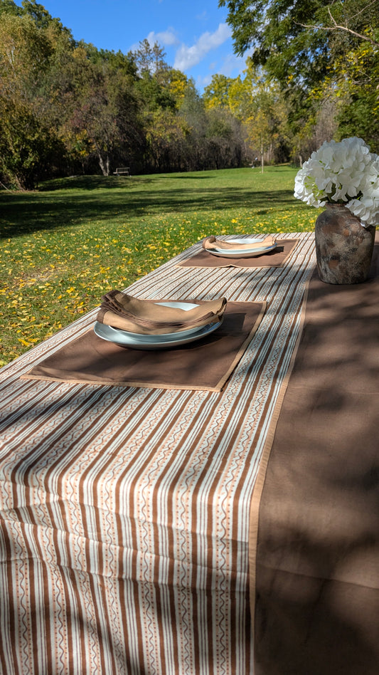 Outdoor dining table setup with a brown and cream striped table runner, matching placemats, plates, folded napkins, and a vase of white flowers in a grassy park setting.