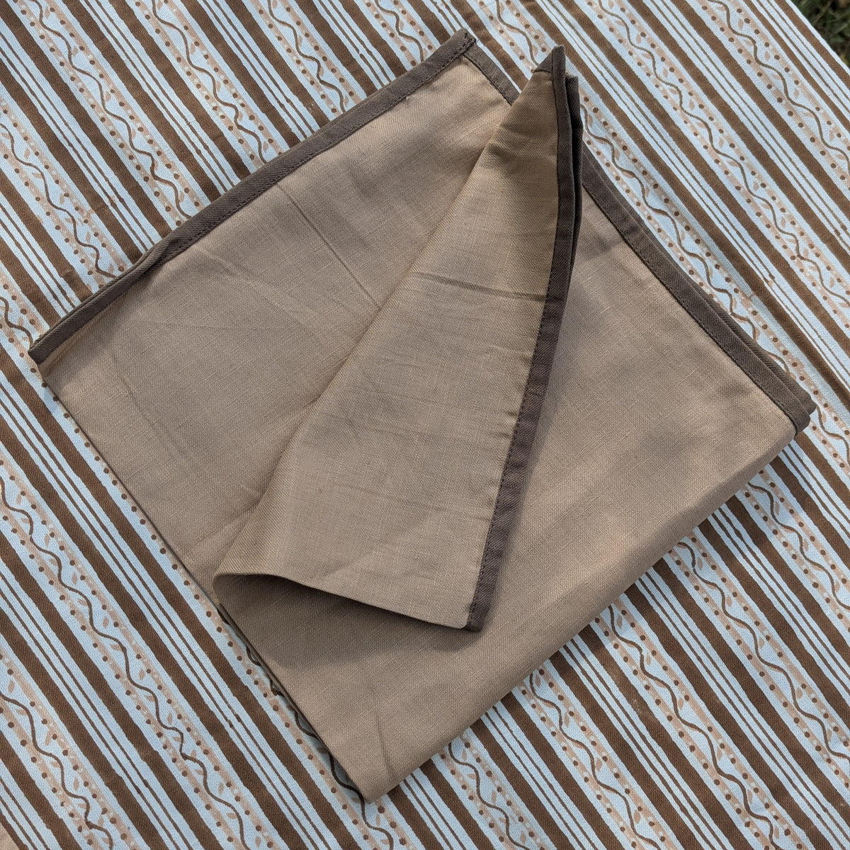 A folded dusty pink fabric item, likely a scarf, shawl, or napkin, with dark brown border trim, displayed on a brown and white patterned striped textile background.