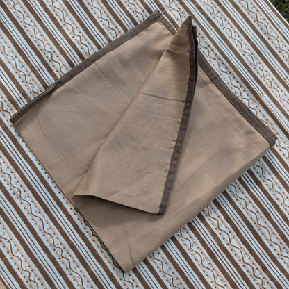 A folded dusty pink fabric item, likely a scarf, shawl, or napkin, with dark brown border trim, displayed on a brown and white patterned striped textile background.