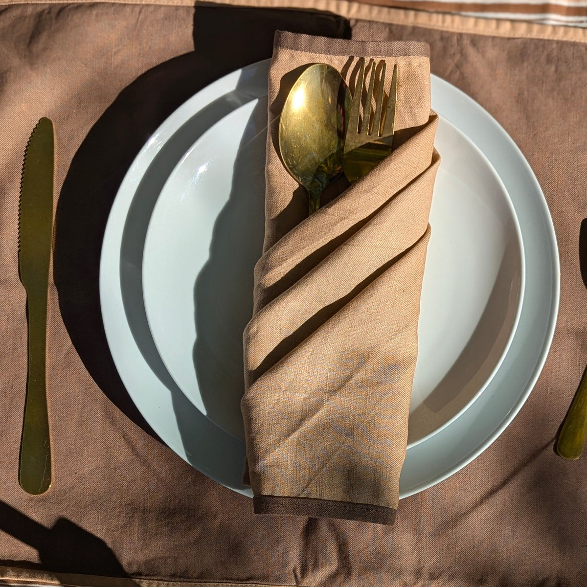 A styled table setting featuring a brown linen napkin folded around gold-toned cutlery, placed on a white plate over a brown placemat with a matching knife and cutlery visible.
