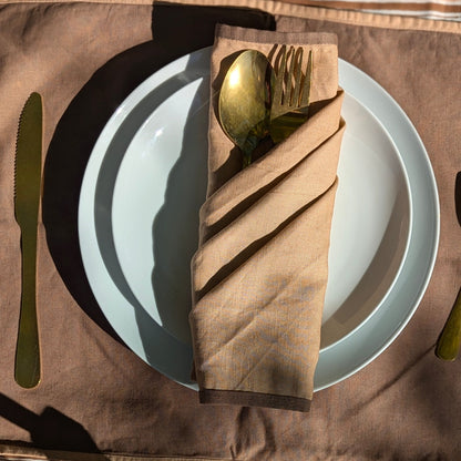 A styled table setting featuring a brown linen napkin folded around gold-toned cutlery, placed on a white plate over a brown placemat with a matching knife and cutlery visible.