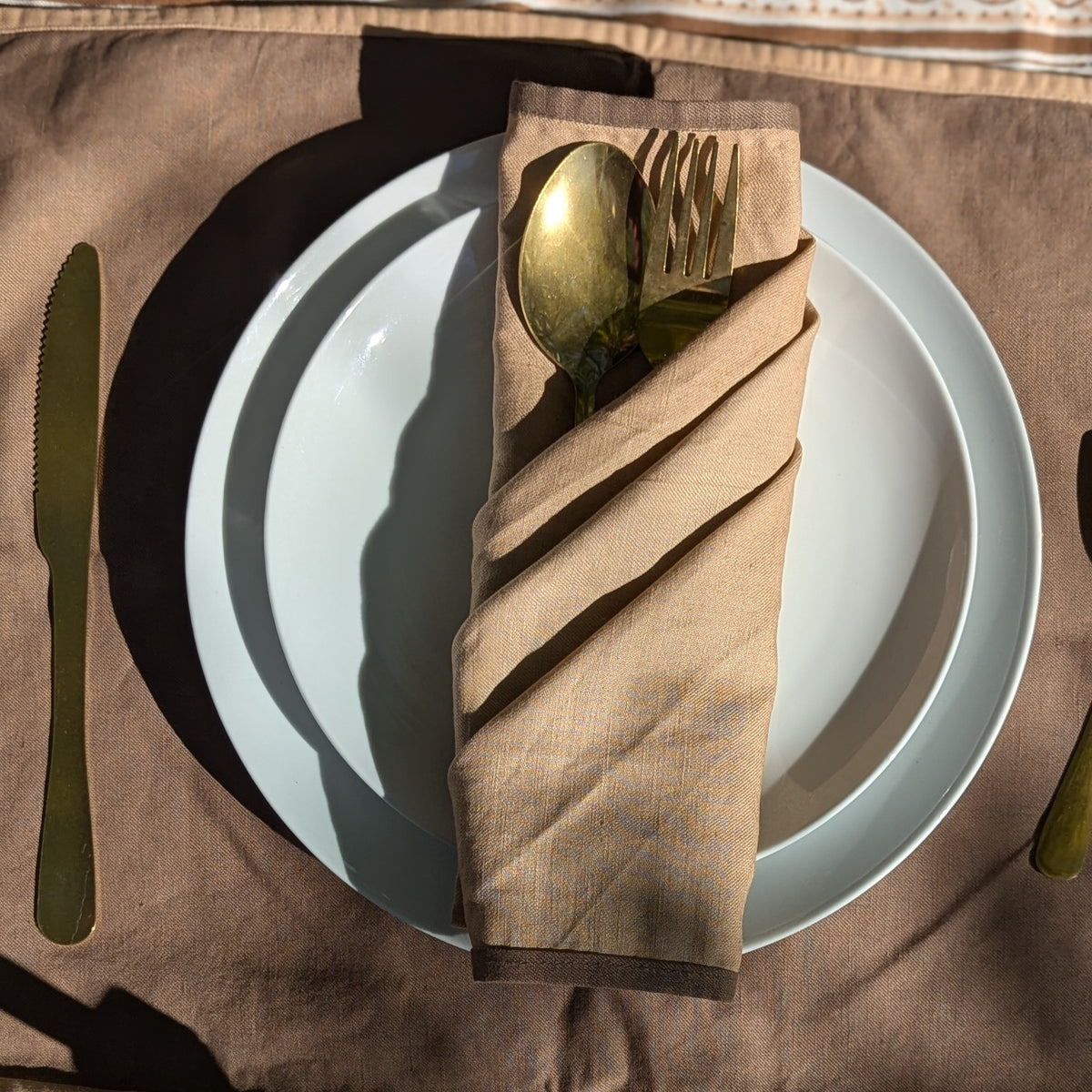 A folded beige linen napkin with a gold spoon and fork tucked inside, arranged on a white plate over a brown tablecloth.