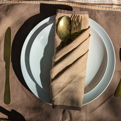 A folded beige linen napkin with a gold spoon and fork tucked inside, arranged on a white plate over a brown tablecloth.