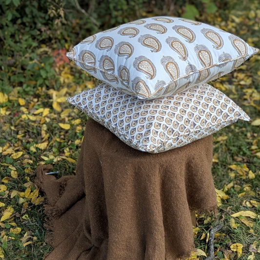 Two decorative throw pillows with a beige and brown paisley pattern are stacked on top of a brown knit blanket outdoors on grass with scattered yellow leaves.
