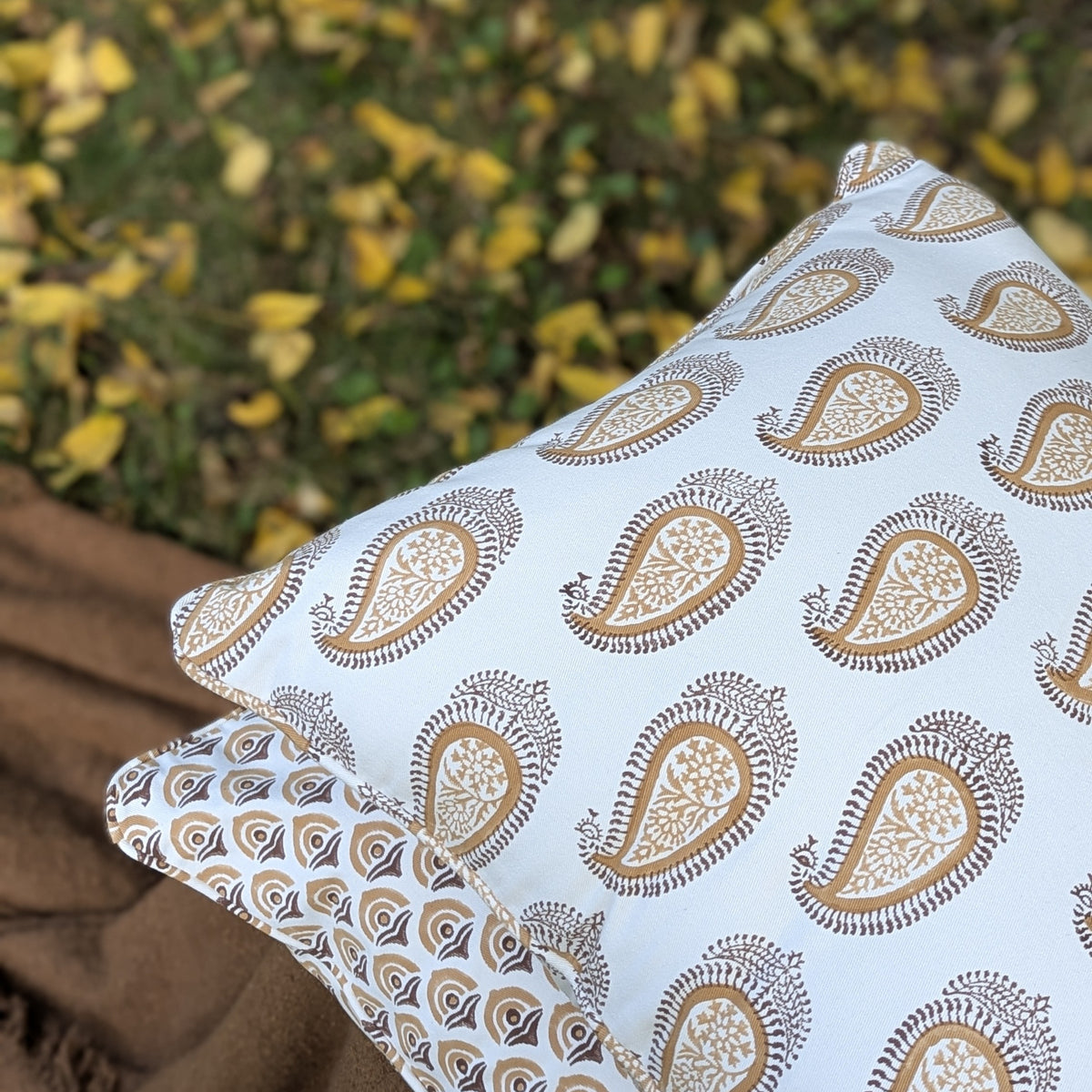 A square decorative throw pillow with a white background and a brown paisley pattern, resting on a brown blanket with a blurred background of green grass and yellow autumn leaves.