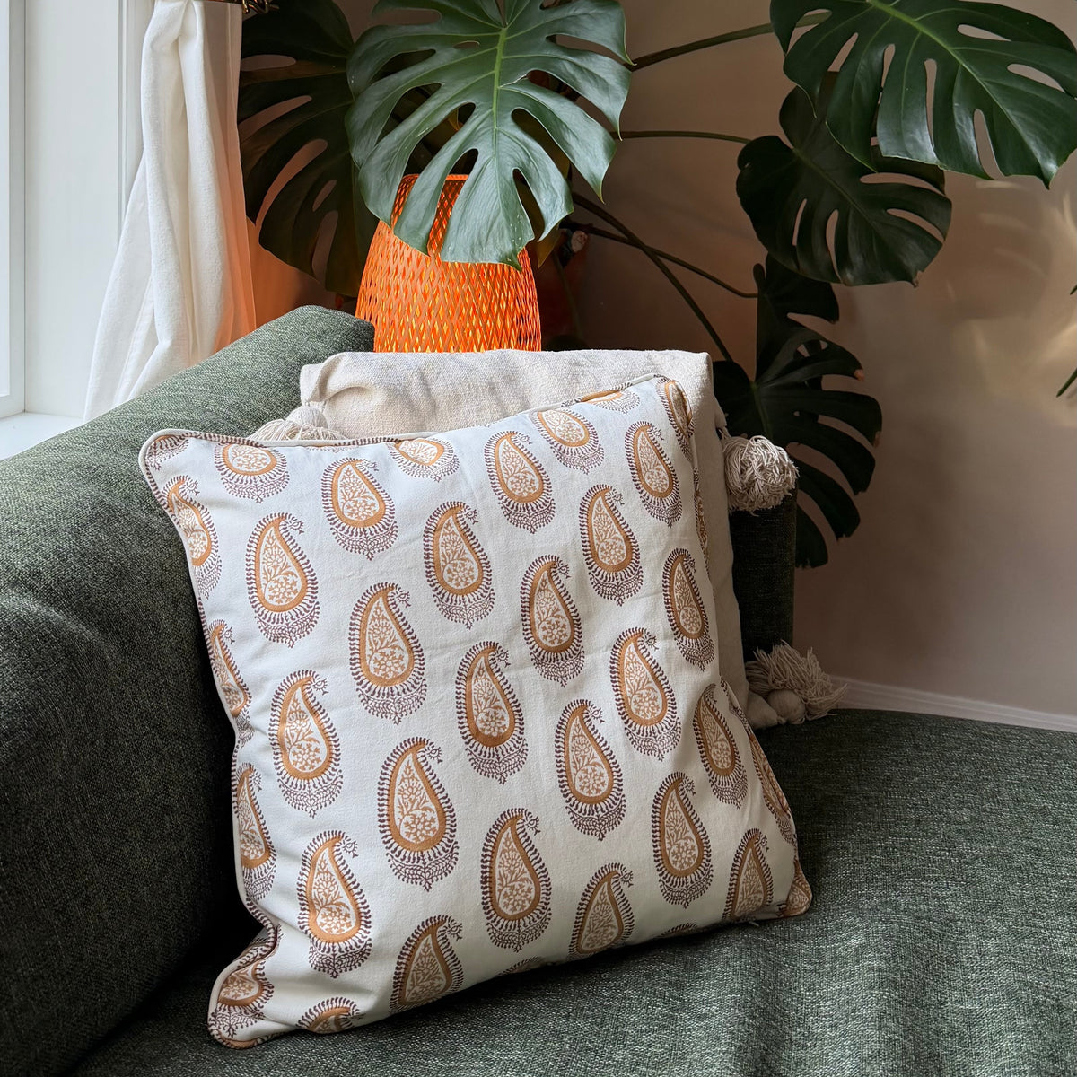A square throw pillow with a beige and brown paisley pattern resting on a green sofa next to a large potted Monstera plant.