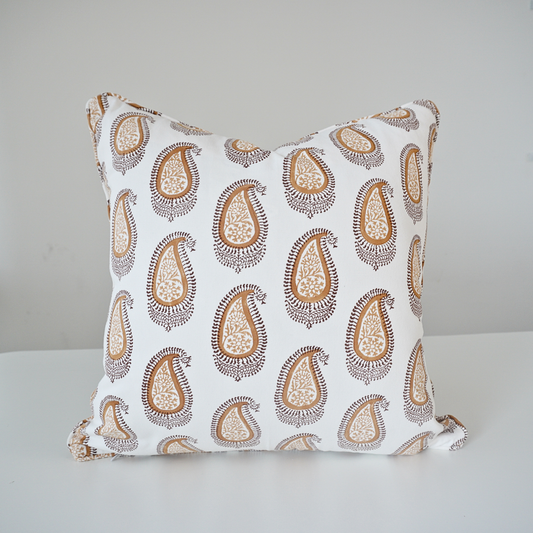 A square throw pillow with a white background and a repeating brown and beige paisley pattern, resting on a white surface against a light gray wall.