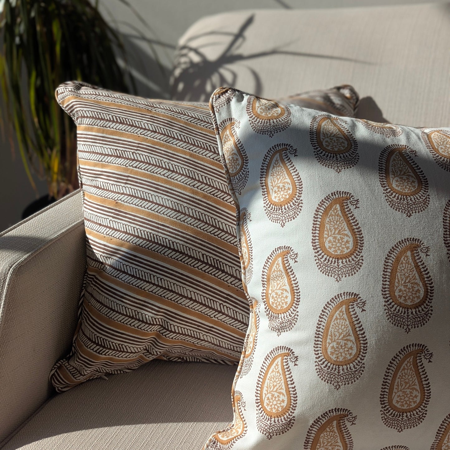 A close-up of two handcrafted cushion covers styled on a sofa in natural light. The cushion on the left features the diagonal striped Serere Soil design in warm earthy tones, paired with a right-hand cushion displaying an intricate brown paisley pattern on a white base. This combination enhances a cozy, modern rustic Canadian farmhouse aesthetic.