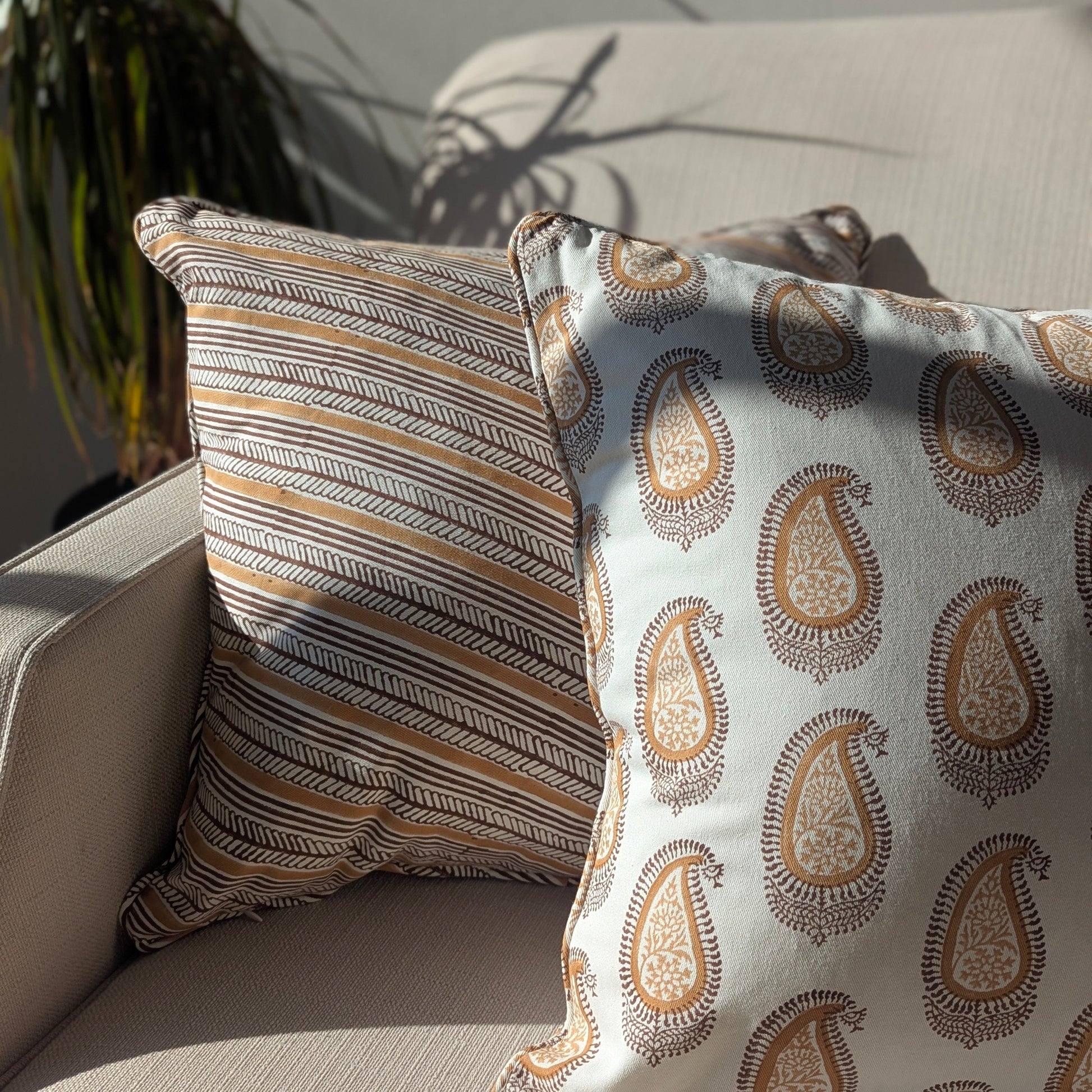 A close-up of two handcrafted cushion covers styled on a sofa in natural light. The cushion on the left features the diagonal striped Serere Soil design in warm earthy tones, paired with a right-hand cushion displaying an intricate brown paisley pattern on a white base. This combination enhances a cozy, modern rustic Canadian farmhouse aesthetic.