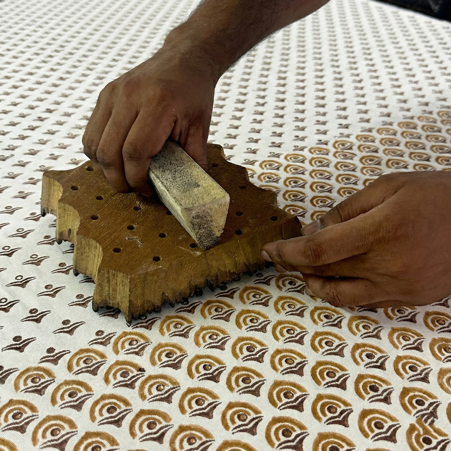 A close-up of an artisan's hands pressing a carved wooden block onto cream fabric, stamping a warm ochre and brown geometric arch pattern. This showcases the traditional hand-block printing process for handcrafted textiles, highlighting the authentic, artisanal quality perfect for a cozy modern Canadian farmhouse aesthetic.