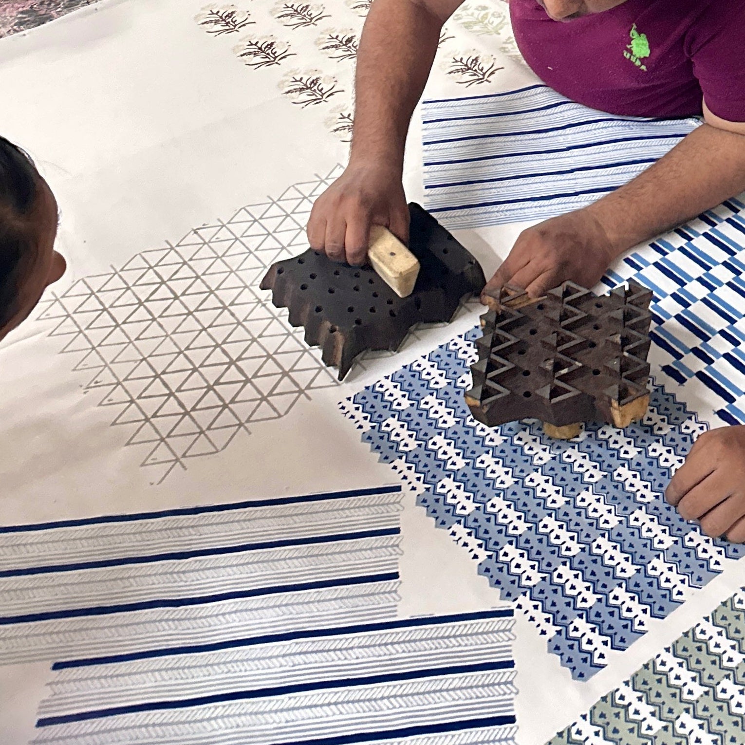 Overhead view of two artisans collaboratively pressing large carved wooden stamps onto a wide expanse of white 100% cotton fabric. The image captures the authentic traditional hand block printing process, showcasing the Moroccan Meadow Mist geometric triangle motif being stamped alongside other intricate blue and green textile patterns. Authentic handmade home decor craftsmanship in action.