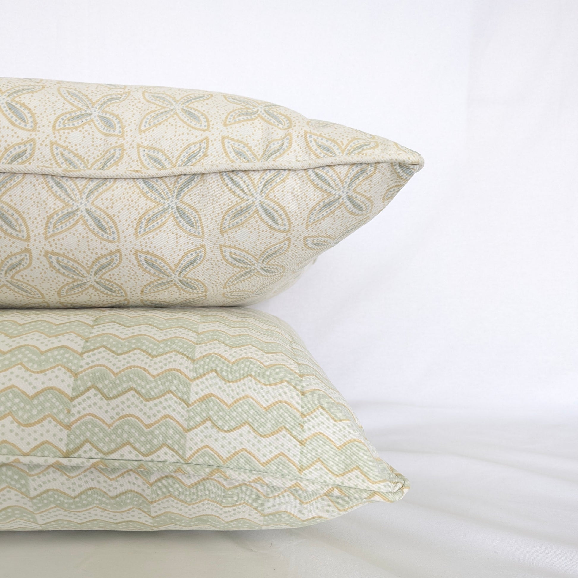 A close-up side view of a stack of two hand-block printed cushions against a crisp white background. The top cushion features the delicate soft blue and tan geometric floral pattern of the Bali plumeria Breeze design, elegantly resting on a coordinating wavy sage green and tan chevron patterned cushion. This detail shot highlights the neatly finished piped edges, showcasing the high quality of these handcrafted textiles that perfectly add a light, cozy modern rustic Canadian farmhouse aesthetic to any home